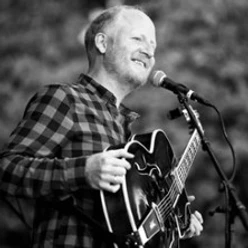 Ein Musiker mit Gitarre steht lächelnd am Mikrofon und spielt auf einer Bühne in schwarzweiß.