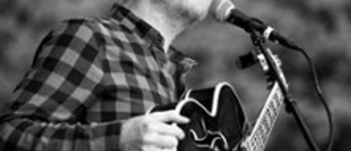 Ein Musiker mit Gitarre steht lächelnd am Mikrofon und spielt auf einer Bühne in schwarzweiß.