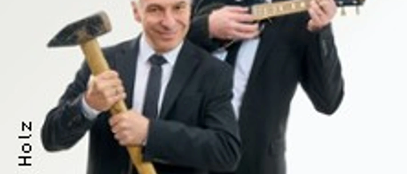 Zwei Männer in Anzügen posieren lachend, einer mit Ukulele, der andere mit Hammer.