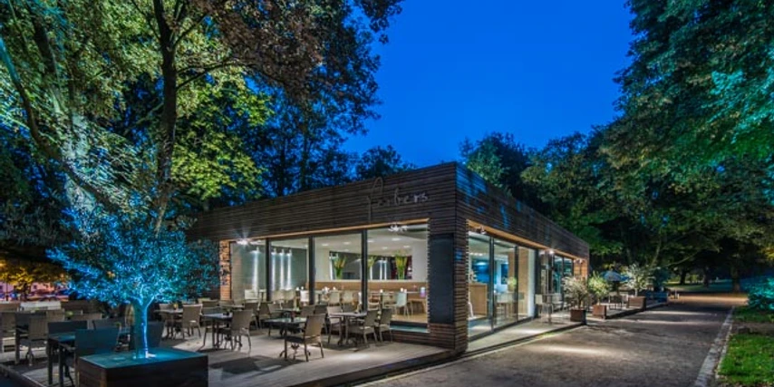 ferbers Außenansicht des Cafés in der Dämmerung mit bestuhlter Außenterrasse und Bäumen ringsherum. Links führt ein Weg vorbei. Drinnen ist es beleuchtet.