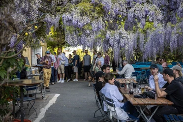 Schwimmbad Rhein-Sommergarten Menschen sitzen und stehen im Biergarten unter herabhängenden lila Blütentrauben