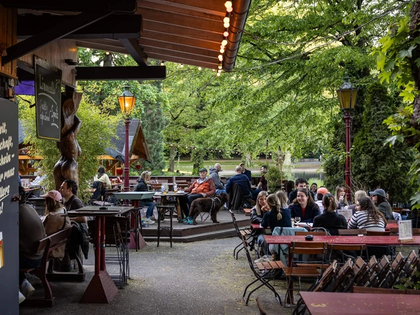 wl-260430-006.jpg Belebter Biergarten im Grünen mit Holztischen, Laternen und Gästen unter Bäumen