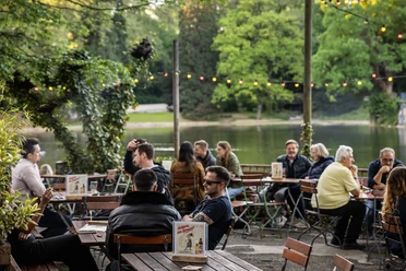 HELLERS Volksgarten Personen sitzen in einer Außengastronomie am Seeufer, umgeben von Grün und Lichterkette.