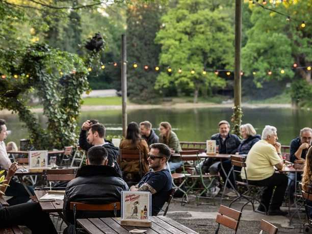 HELLERS Volksgarten People are sitting at an outdoor restaurant on the lakeshore, surrounded by greenery and string lights.