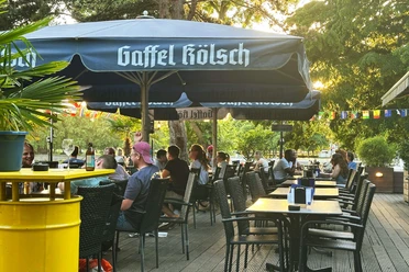 Beer Garden at Aachener Weiher Outdoor dining with guests under large blue umbrellas in the evening light.