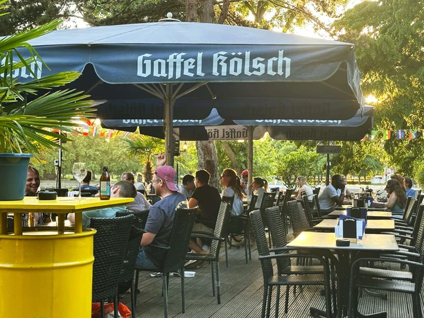 Biergarten am Aachener Weiher Außengastronomie mit Gästen unter großen blauen Sonnenschirmen im Abendlicht.