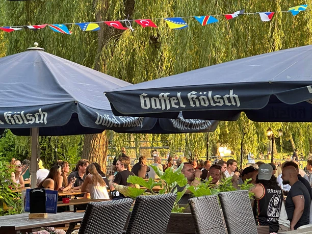 Biergarten am Aachener Weiher Menschen sitzen im Biergarten unter großen blauen Sonnenschirmen bei Grün und Abendlicht