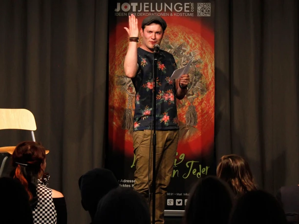 Veranstaltungsreihe „Sperenzien“ von Jot Jelunge Person auf kleiner Bühne spricht ins Mikro und gestikuliert vor Publikum