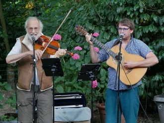 Klaus der Geiger & Marius Peters Zwei Musiker spielen Geige und Gitarre im Garten vor Mikrofonen und blühenden Sträuchern