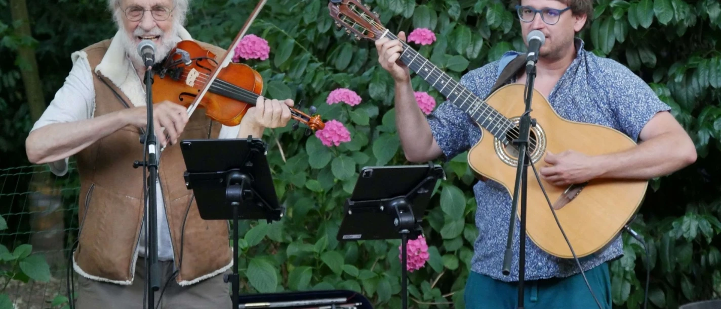 Klaus der Geiger & Marius Peters Zwei Musiker spielen Geige und Gitarre im Garten vor Mikrofonen und blühenden Sträuchern