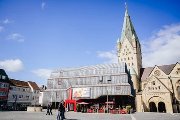 Diözesanmuseum Außen neuer Banner.jpg Das Diözesanmuseum Paderborn von vorne. ES ist ein schöner Tag mit blauem Himmel. Das Außenbanner zeigt die Ausstellungsgrafik von Die Hände zum Himmel