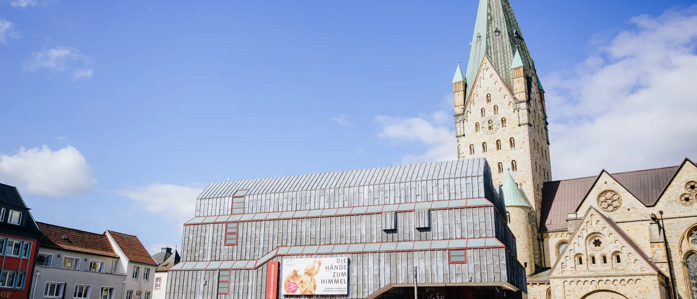 Diözesanmuseum Außen neuer Banner.jpg Das Diözesanmuseum Paderborn von vorne. ES ist ein schöner Tag mit blauem Himmel. Das Außenbanner zeigt die Ausstellungsgrafik von Die Hände zum Himmel