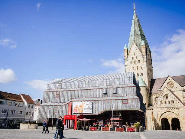 Diözesanmuseum Außen neuer Banner.jpg Das Diözesanmuseum Paderborn von vorne. ES ist ein schöner Tag mit blauem Himmel. Das Außenbanner zeigt die Ausstellungsgrafik von Die Hände zum Himmel