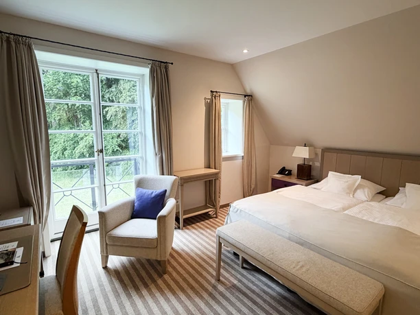 Hotelværelser på skovhuset Heiligenberg Lyst hotelværelse med dobbeltseng, lænestol, skrivebord og udsigt til grønne omgivelser.