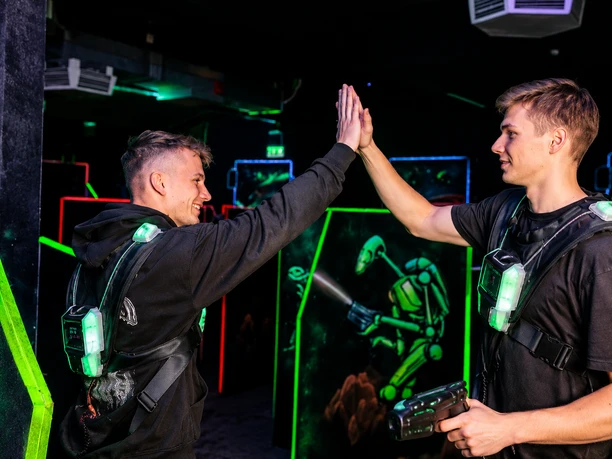 Laser Heroe (c) Laser Heroes - Lasertag Bremerhaven.png Zwei junge Männer geben sich beim Lasertag in einer neonbeleuchteten Arena ein High-Five.