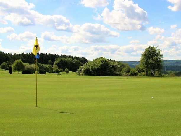 Golfanlage Gimborner Land Weite Golfanlage mit Flagge und Ball vor bewaldetem Hintergrund unter blauem Himmel.