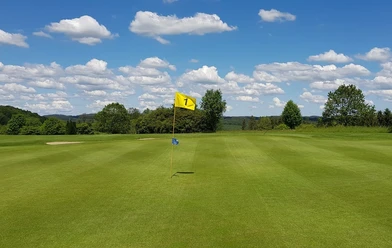 Golfanlage Gimborner Land Weitläufiges Golfgrün mit Fahne der Bahn 1 vor bewaldeter Landschaft und blauem Himmel
