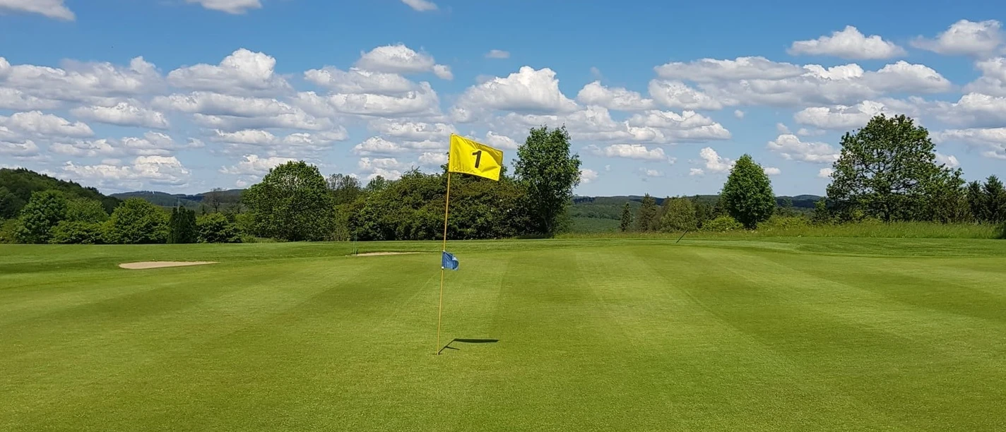 Golfanlage Gimborner Land Weitläufiges Golfgrün mit Fahne der Bahn 1 vor bewaldeter Landschaft und blauem Himmel