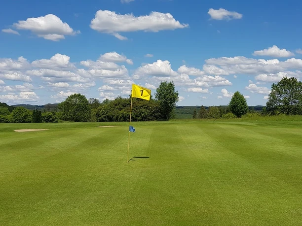 Golfanlage Gimborner Land Weitläufiges Golfgrün mit Fahne der Bahn 1 vor bewaldeter Landschaft und blauem Himmel