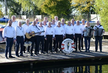 Shanty-Chor-Wunstorf-2026.jpg Der Wunstorfer Shanty Chor posiert in Matrosenkleidung mit Instrumenten auf einem Holzsteg am Wasser.
