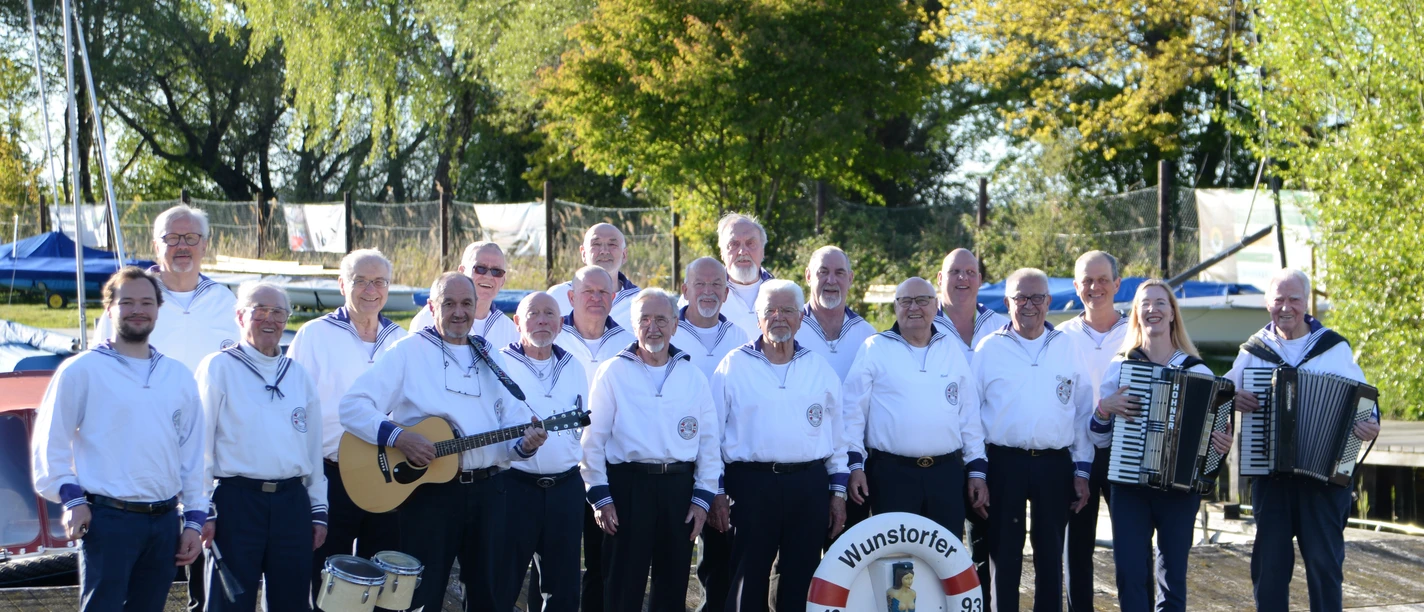 Shanty-Chor-Wunstorf-2026.jpg Der Wunstorfer Shanty Chor posiert in Matrosenkleidung mit Instrumenten auf einem Holzsteg am Wasser.