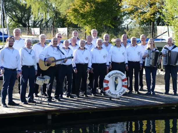 Shanty-Chor-Wunstorf-2026.jpg Der Wunstorfer Shanty Chor posiert in Matrosenkleidung mit Instrumenten auf einem Holzsteg am Wasser.