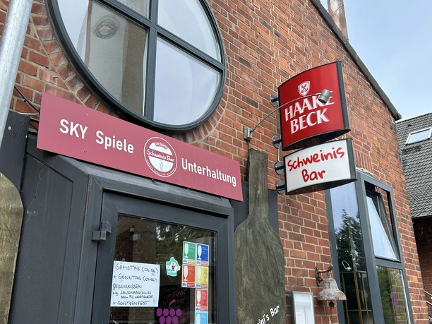 Außenansicht von Schweinis Bar mit Schildern vor der Backsteinfassade und einem runden Fenster.