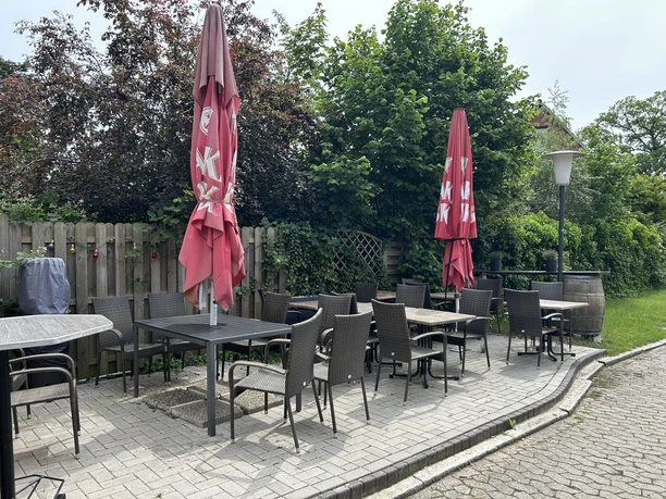 Die gepflasterte Außenterrasse mit Tischen, Stühlen und zwei geschlossenen roten Sonnenschirmen.