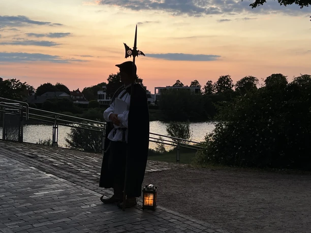 Nienburger Nachtwächter Ein Nachtwächter in historischer Tracht mit Hellebarde und Laterne am Ufer bei Sonnenuntergang.