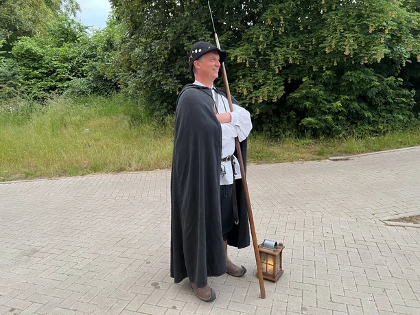 Ein Nachtwächter in historischer Kleidung mit schwarzem Umhang, Speer und einer leuchtenden Laterne.
