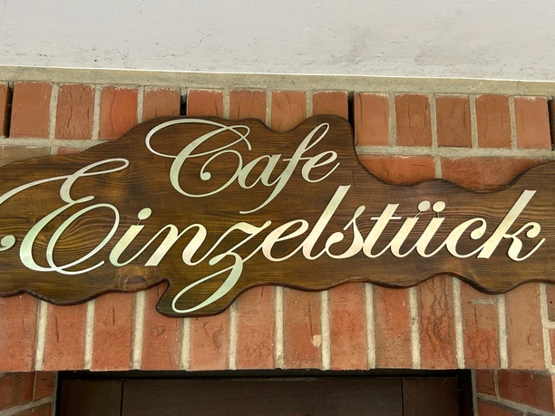Eingang Café Einzelstück Fischförmiges Holzschild mit dem Schriftzug „Cafe Einzelstück“ an einer Backsteinwand über dem Eingang.