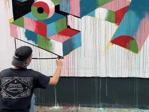 Eine Person arbeitet kniend an einem farbintensiven geometrischen Wandbild mit Tropfspuren.
