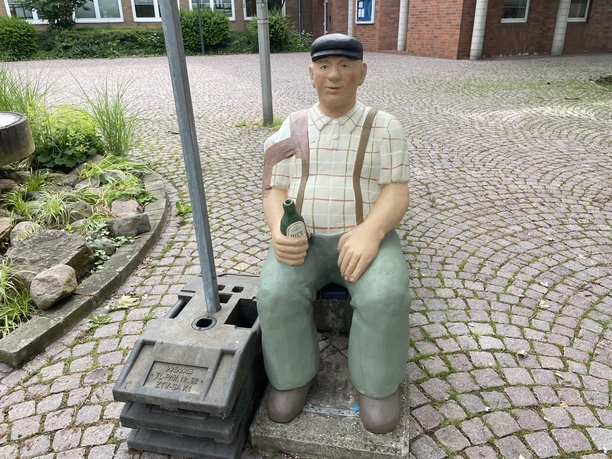 Herr Hagen Lebensgroße Skulptur 'Herr Hagen', sitzend mit Schiebermütze und Bierflasche auf einer Bank.