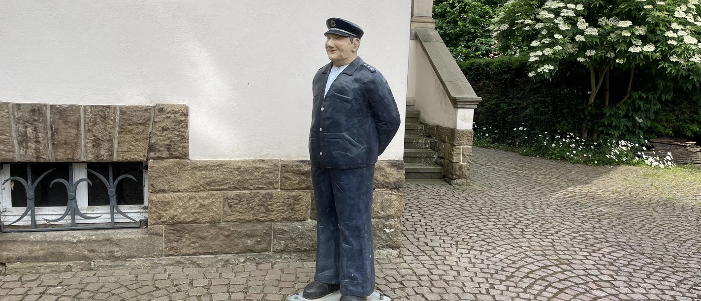 Lebensgroße Skulptur des Wachtmeisters Schulze in Uniform vor einem hellen Haus auf Kopfsteinpflaster.
