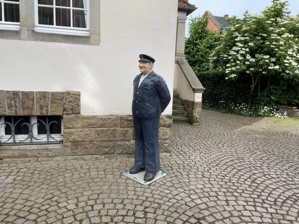 Wachmeister Schulze Lebensgroße Skulptur des Wachtmeisters Schulze in Uniform vor einem hellen Haus auf Kopfsteinpflaster.