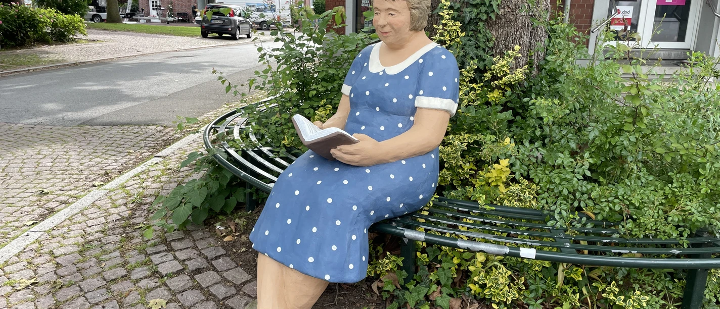 Hermine Die Skulptur Hermine sitzt in einem blauen, gepunkteten Kleid lesend auf einer grünen Parkbank.