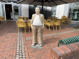 Anzeigen Peter Lebensgroße Skulptur 'Anzeigen Peter' steht auf einem gepflasterten Platz vor einem Café.