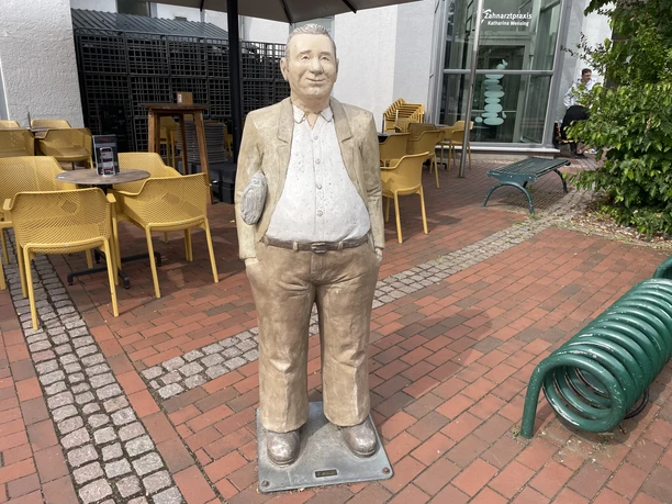 Anzeigen Peter Lebensgroße Skulptur „Anzeigen Peter“ auf einem gepflasterten Platz vor einer Eisdiele in Petershagen.