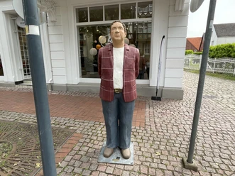 Herr Meyer Lebensgroße Skulptur "Herr Meyer" mit rot karierter Jacke steht auf einem Kopfsteinpflaster-Gehweg.