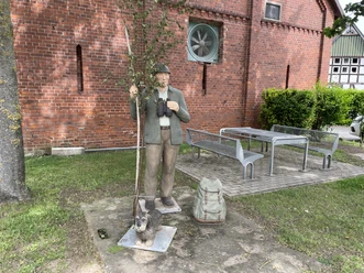 Lebensgroße Figur des Jägers Ulli mit seinem Dackel Anton vor einer roten Ziegelmauer.