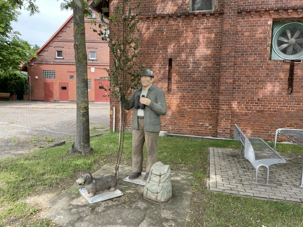 Figur des Jägers Ulli mit Dackel Anton, Fernglas und Rucksack vor einem Backsteingebäude.