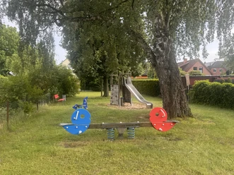 Spielplatz Jahnstraße Wippe auf einer Grünfläche, im Hintergrund ist ein großer Baum zu sehen und dahinter eine Rutsche. Links an der Seite steht eine rote Bank.