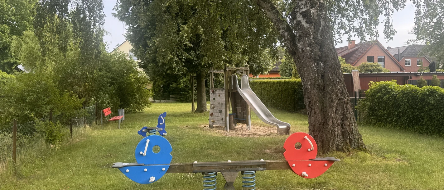 Spielplatz Jahnstraße Wippe auf einer Grünfläche, im Hintergrund ist ein großer Baum zu sehen und dahinter eine Rutsche. Links an der Seite steht eine rote Bank.