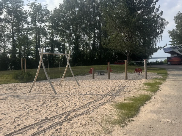 Das Bild zeigt einen Spielplatz mit Sandbereich. Darauf steht eine Schaukel, ein Klettergerüst mit Reckstangen in verschiedenen Höhen und einzelne Wippspielzeuge.
