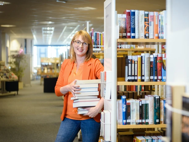 23-06-06_BundesprogrammZIZ Stadtbibliothek_ Anika Schmidt (c) Antje Schimanke_WEB.jpg Eine lächelnde Mitarbeiterin der Bibliothek hält einen Stapel Bücher neben einem Bücherregal.