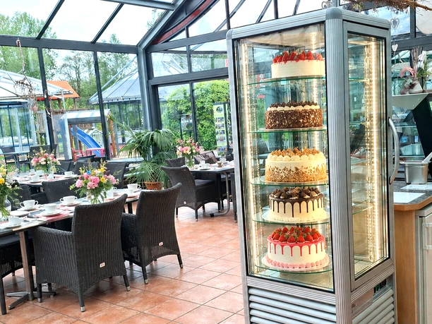 Cafe mit Tortenturm.png Eine Tortenvitrine in einem hellen Café mit gedeckten Tischen und Blick auf einen Spielplatz.