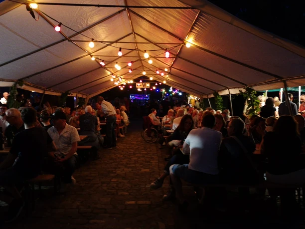 Impression Weinfest 2024.jpeg Gäste des Weinfests Hengstforde feiern abends unter Lichterketten in einem langen Festzelt.