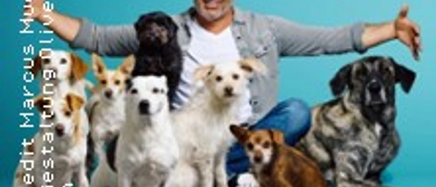 Andreas Ohligschläger lächelt mit mehreren Hunden vor türkisfarbenem Hintergrund