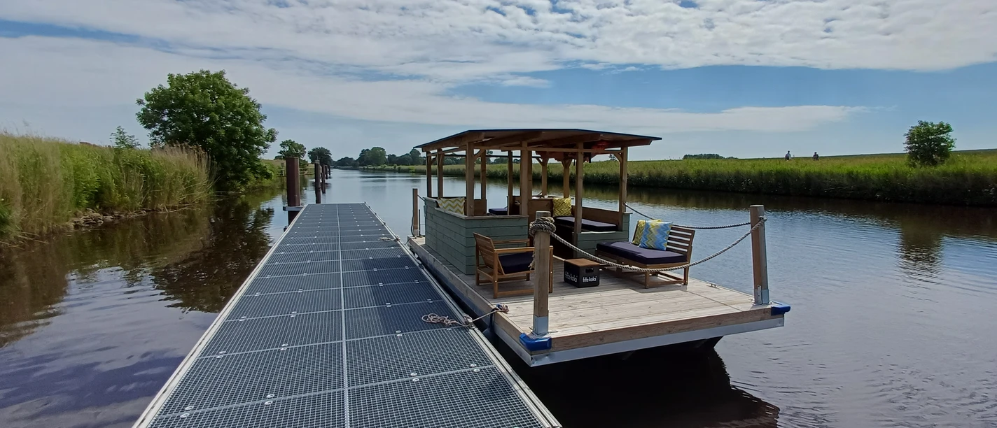 NoBoatGermany Nordseebad Otterndorf Ein überdachtes Floß mit Sitzgelegenheiten am Steg eines Kanals unter sommerlichem Himmel.
