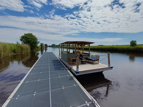 NoBoatGermany Nordseebad Otterndorf Ein überdachtes Floß mit Sitzgelegenheiten am Steg eines Kanals unter sommerlichem Himmel.